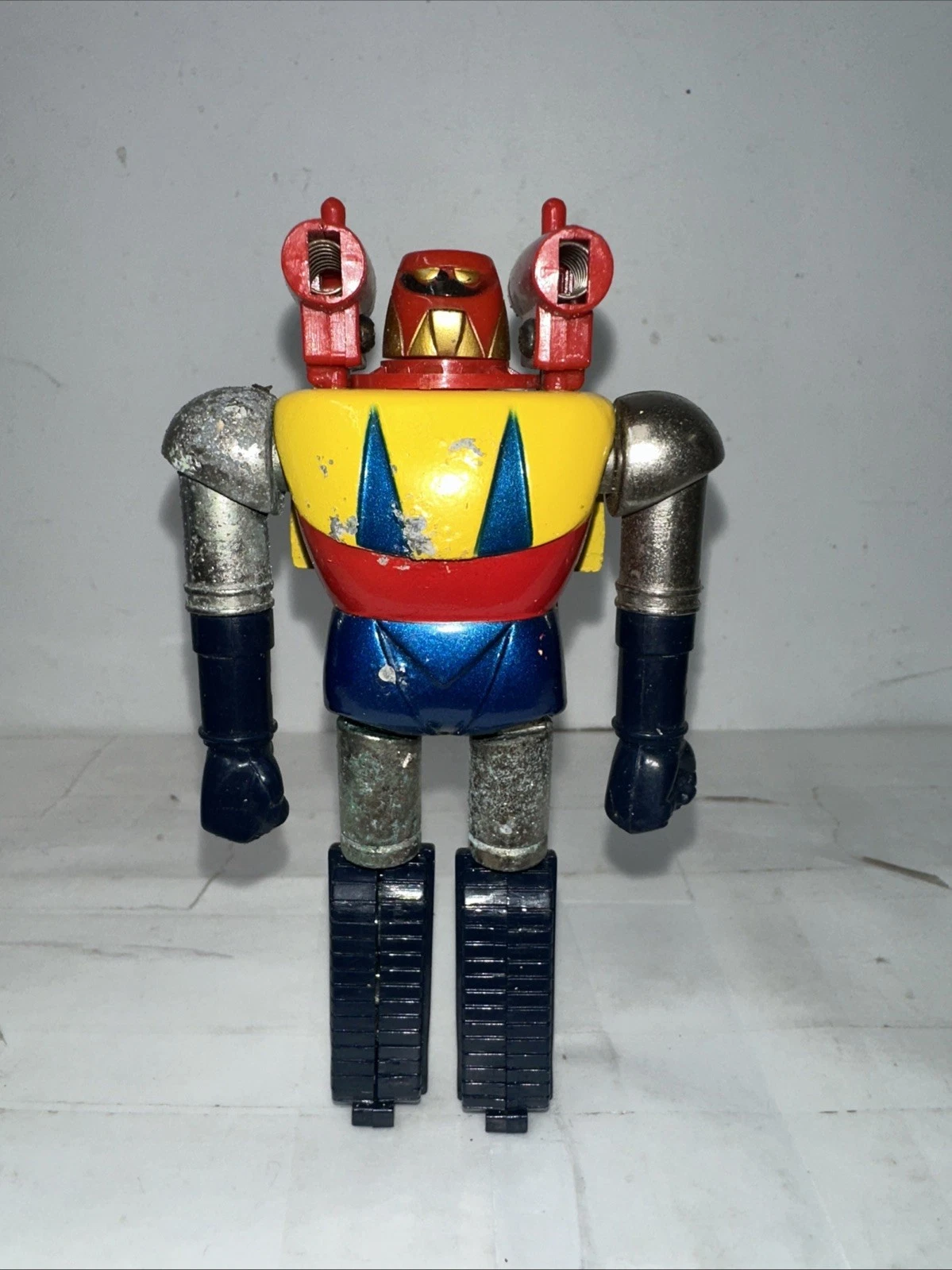 A small die-cast Japanese robot toy: red head with a gold faceplate, yellow chest with two blue triangles, chrome shoulders, dark blue arms, and tank treads for feet. Two red rocket pods are mounted on the shoulders. The paint is chipped from play.