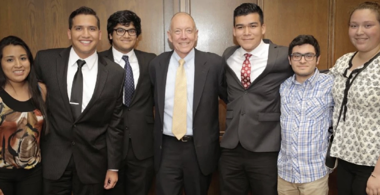 Six college students in suits and dress clothes standing shoulder to shoulder with Ted Mitchell, then U.S. Under Secretary of Education, in a wood-paneled room. Everyone is smiling.